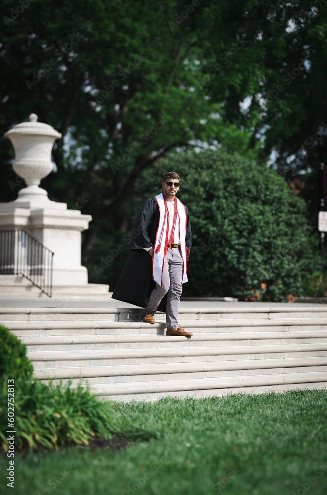 Fototapeta premium University of Louisville male graduate walking down the stairs of one of the campus buildings wearing a gown and honor ropes