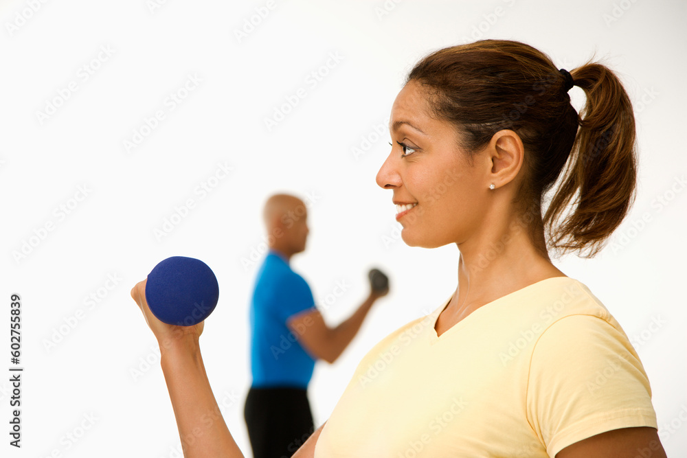 Close up of smiling mid adult multiethnic woman exercising using dumbbells with mid adult multiethnic man in background. Close up of smiling mid adult multiethnic woman exercising using dumbbells with mid adult multiethnic man in background.