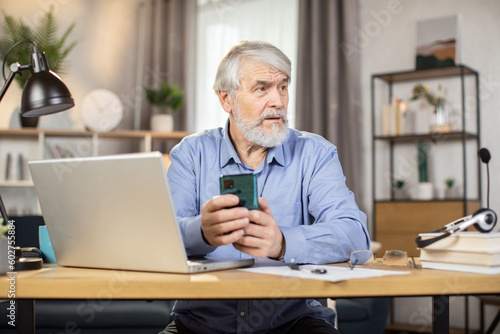 Focused elderly adult in headset and glasses holding smartphone while sitting at desk with portable computer on it. Efficient employee texting short message in online chat via app in home office.