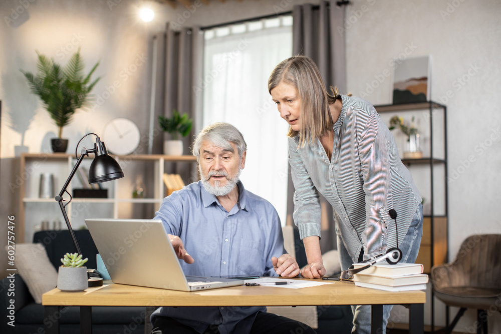 Attractive female standing with pen in hand while senior male looking through paper document on desk near laptop. Experienced entrepreneurs correcting written contract together in home office interior Attractive female standing with pen in hand while senior male looking through paper document on desk near laptop. Experienced entrepreneurs correcting written contract together in home office interior