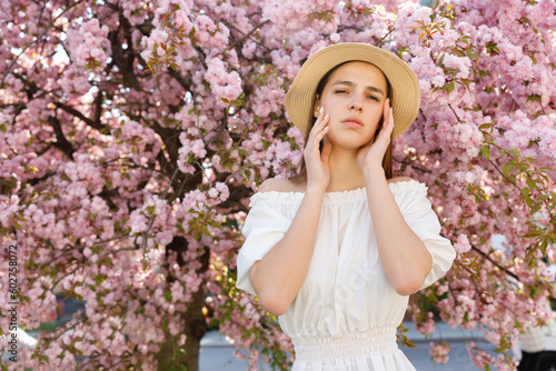 Charming Cute Caucasian Dark Haired Lady in Straw Hat Standing in the Park, Posing with Hands Beauty Poses, Near Sakura Tree With Pink Flowering. Femininity and Natural Beauty Concept