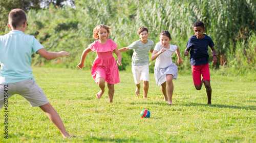 International group of active tweenagers having fun together outdoors, playing football on green grass in summertime