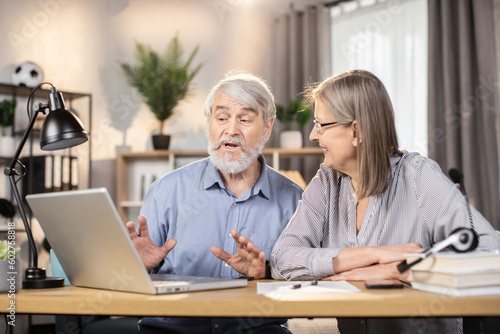 Elegant senior lady in glasses and gray-haired elderly man utilizing portable computer for distant work at home. Efficient freelancers studying details of temporary contract on digital screen.