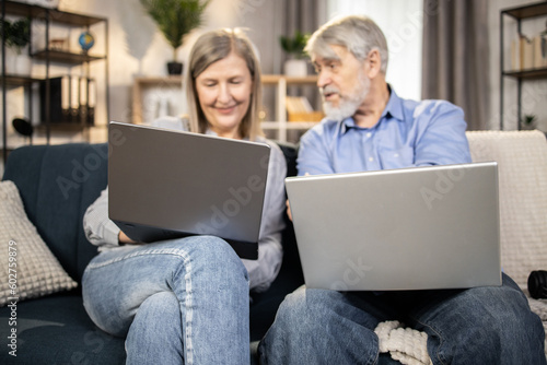 Gray-haired male in mature age reaching out hand to computer held by attractive senior lady sitting on couch in apartment. Married couple in jeans browsing online fashion store via internet app. photo