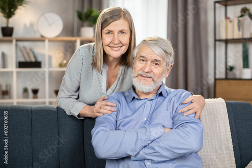 Portrait of loving family man and wife in casual wear spending time together in modern studio apartment. Cheerful lady of middle age hugging warmly happy man while standing behind sofa in lounge.