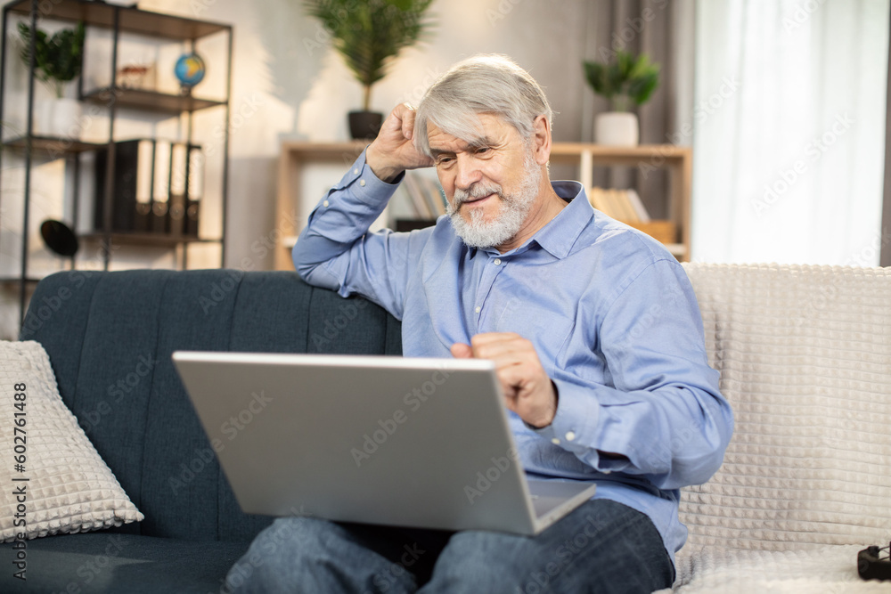 Thoughtful mature man in casual clothes with laptop on knees being engaged in pleasant pastime indoors. Focused caucasian man around 65 watching adventure film online via internet connection at home. Thoughtful mature man in casual clothes with laptop on knees being engaged in pleasant pastime indoors. Focused caucasian man around 65 watching adventure film online via internet connection at home.