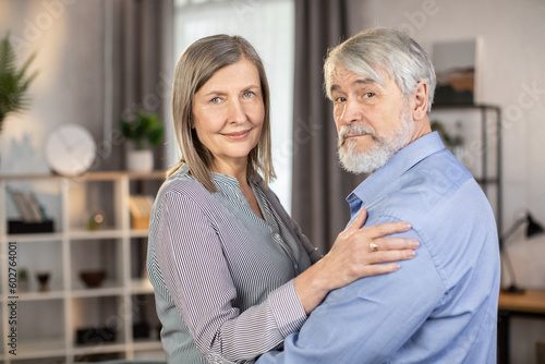Portrait of happy senior married couple looking at camera with tenderness and love while standing in living room. Adoring husband touching shoulder of wife gently. Concept of family treasures.