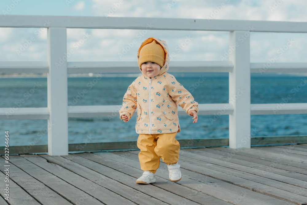 A toddler in a yellow jacket and pants walks on the pier