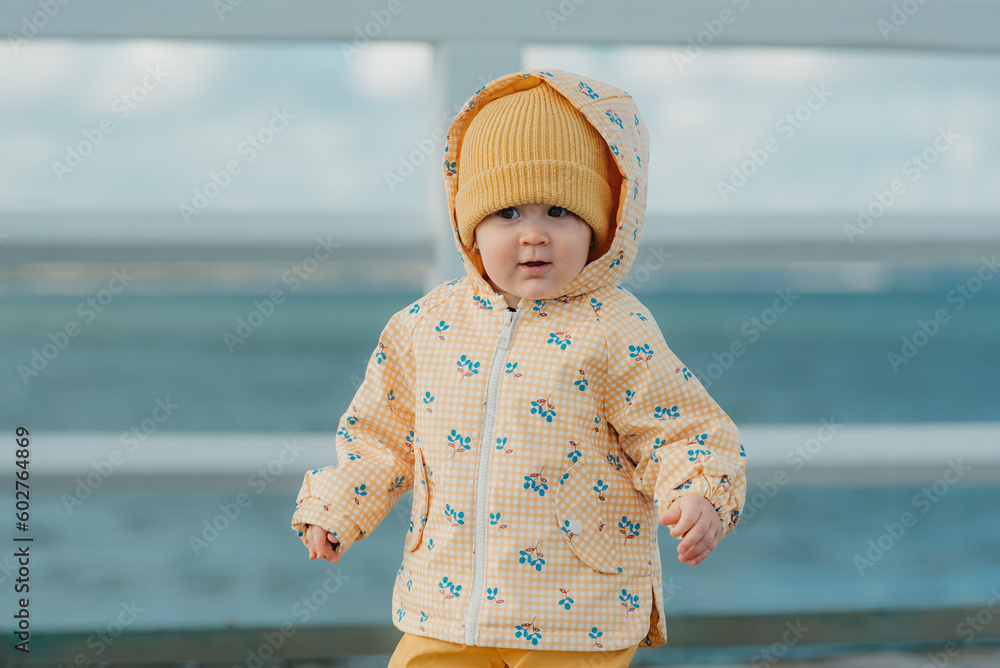 A toddler in a yellow jacket and pants walks on the pier