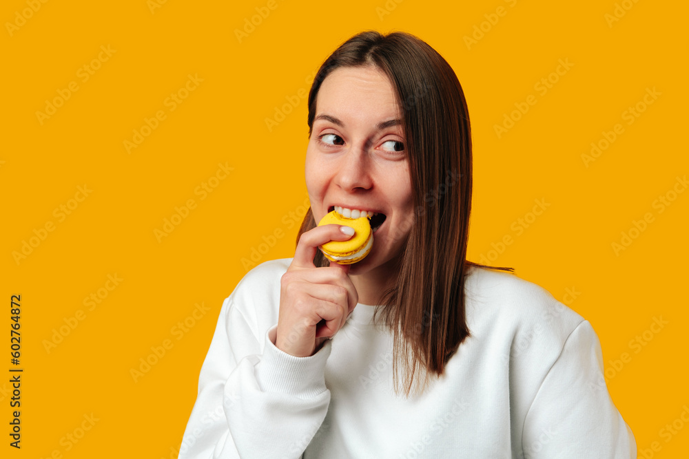 Studio portrait of a playful young woman taking a bite of a yellow macaroon over yellow background. Studio portrait of a playful young woman taking a bite of a yellow macaroon over yellow background.