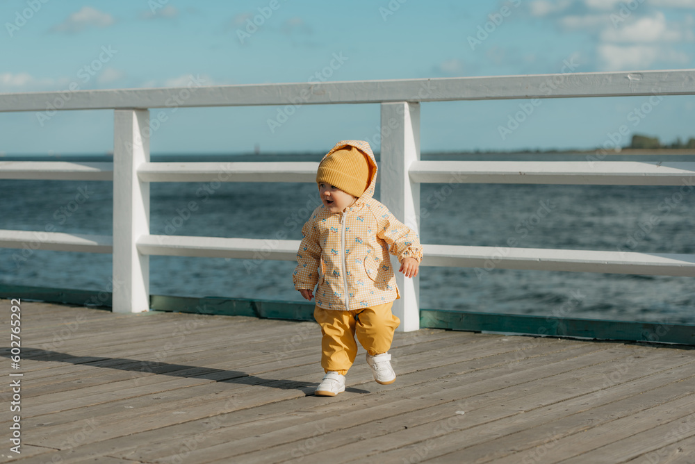 A toddler in a yellow jacket and pants walks on the pier