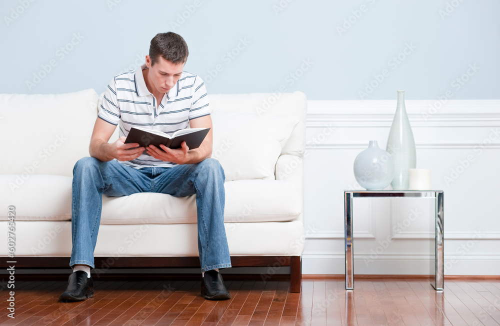 Man sitting and reading on a white couch in his living room. Horizontal format.