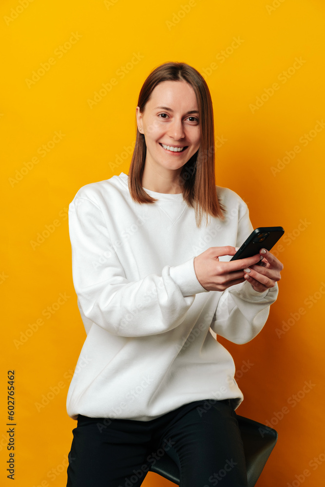 Vertical portrait of a young woman sitting on a chair and holding her phone over yellow background. Vertical portrait of a young woman sitting on a chair and holding her phone over yellow background.