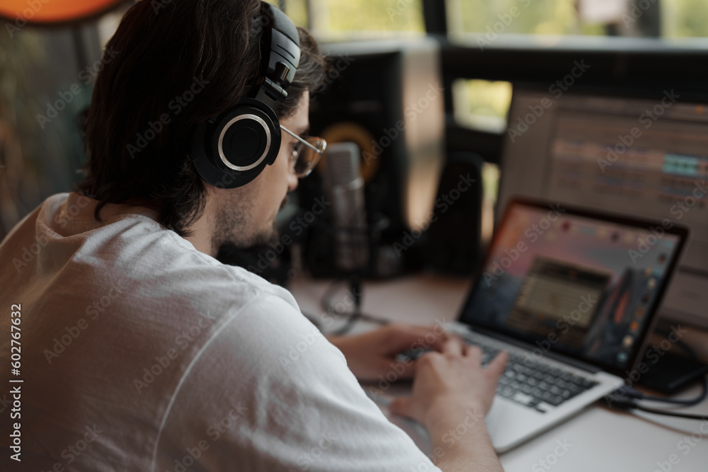 Side view of focused male musician in white t shirt and headphones typing on laptop while recording podcast in modern studio Side view of focused male musician in white t shirt and headphones typing on laptop while recording podcast in modern studio
