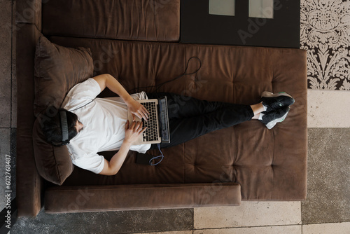 Top view full body of anonymous male musician in white t shirt and headphones lying on comfortable brown sofa and composing music on laptop © arthurhidden