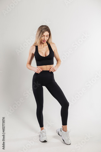 Fashionable pretty fitness woman with blonde hair in fashion black sports outfit with leggings, top and sneakers poses on a white background