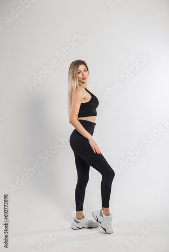 Beautiful young blonde fitness girl in fashionable black sportswear with sneakers, leggings and a top on a white background in the studio