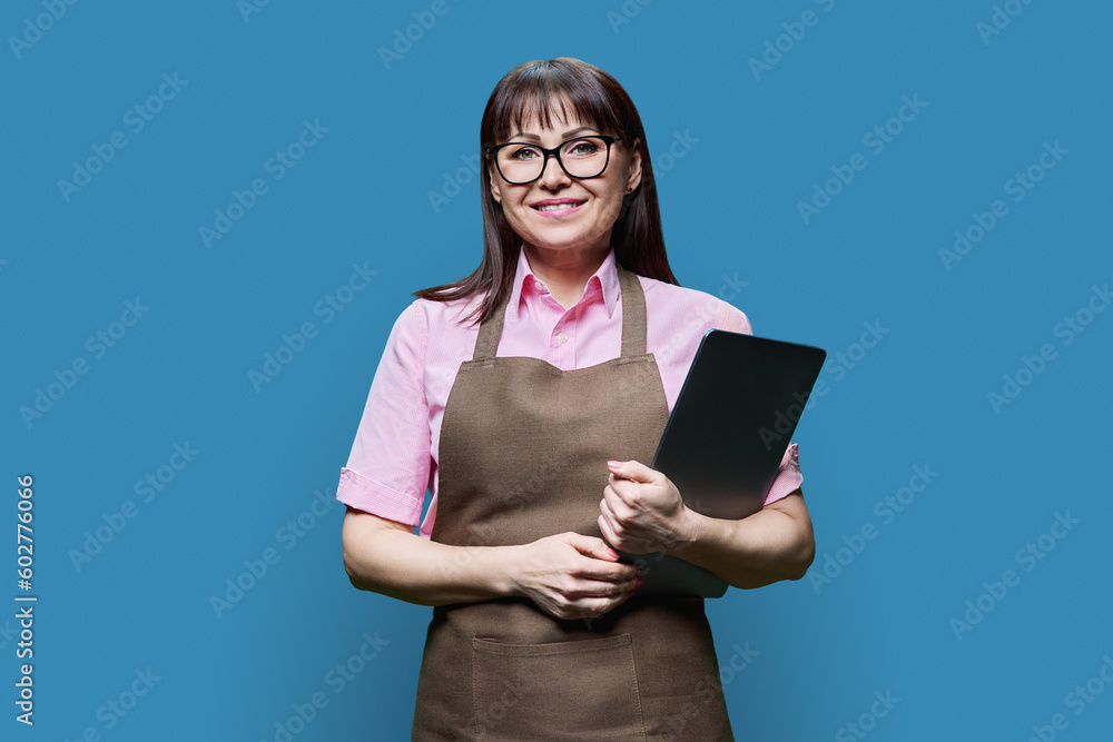 Portrait of middle aged woman in apron with laptop on blue background Portrait of middle aged woman in apron with laptop on blue background