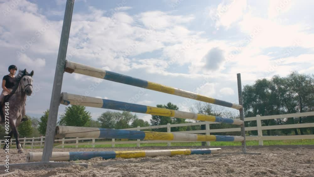 Girl rider on dapple gray horse jumping over triple bars in outdoor arena on a sunny summer day, low angle shot. Horse sports competition concept.