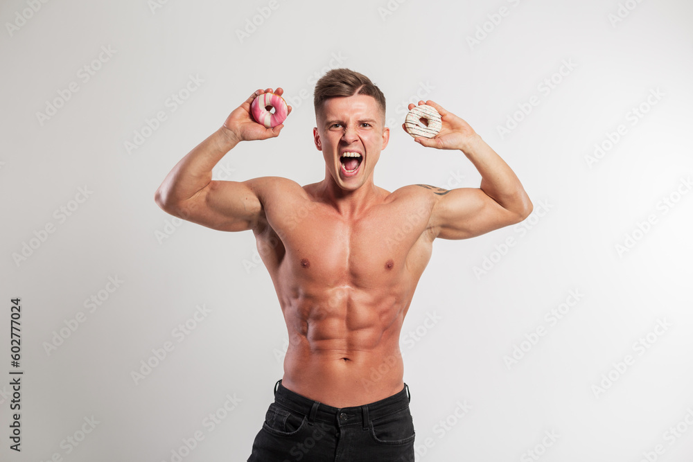 Handsome emotional fitness man with a muscular body screams and holds donuts on a white background. Sport workout and sugar sweet diet, creative idea Handsome emotional fitness man with a muscular body screams and holds donuts on a white background. Sport workout and sugar sweet diet, creative idea