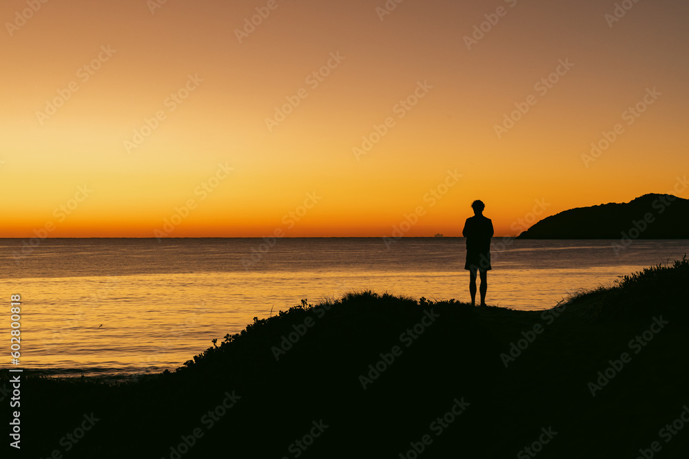 Silhouette of man standing on sand dune overlooking Jimmy's Beach at sunrise. Hawks Nest, NSW Australia