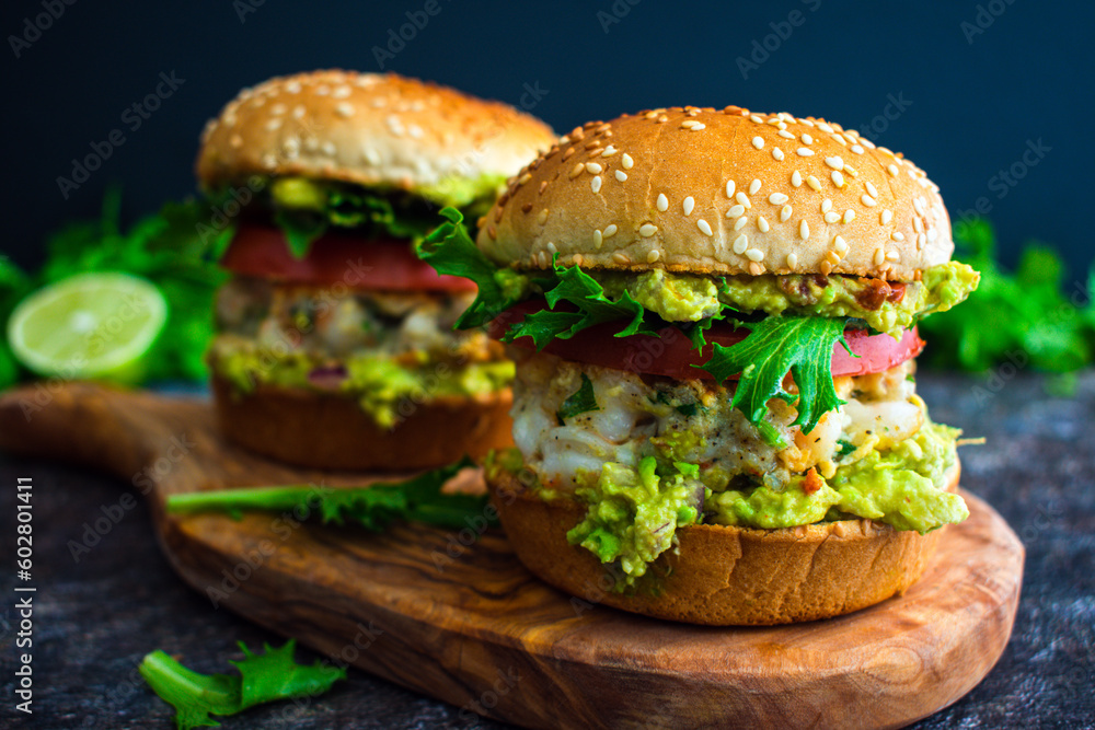 Shrimp Burgers with Cilantro and Guacamole on Sesame Seed Buns: Seafood hamburger sandwiches made topped with guacamole and sliced tomato Shrimp Burgers with Cilantro and Guacamole on Sesame Seed Buns: Seafood hamburger sandwiches made topped with guacamole and sliced tomato