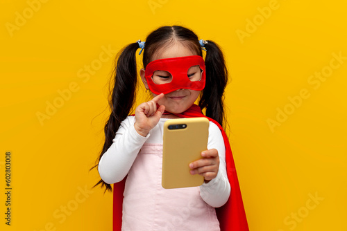 little asian girl in superman costume and mask use smartphone, korean kid in superhero cape typing on phone
