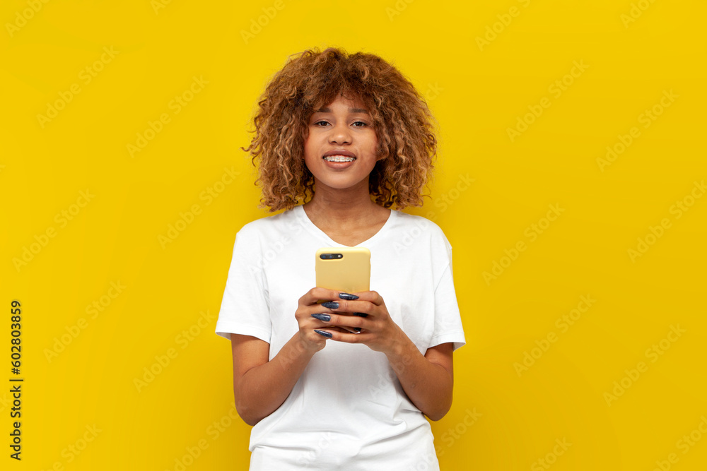 young curly american girl in white t-shirt with braces uses smartphone on yellow isolated background young curly american girl in white t-shirt with braces uses smartphone on yellow isolated background