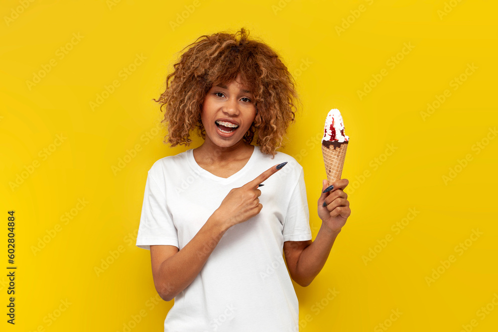 young curly american girl with braces eats sweet ice cream with jam over yellow isolated background young curly american girl with braces eats sweet ice cream with jam over yellow isolated background