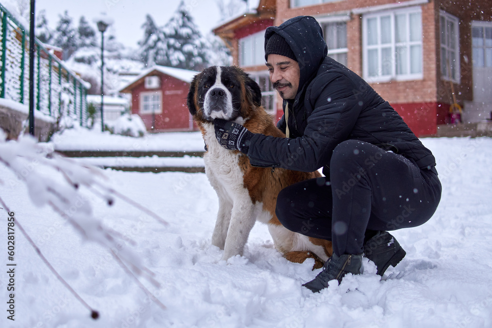 portrait latin man with mustache and hooded jacket, embracing a saint bernard dog together in profile in winter snow in a cabin complex. Horizontal y copy space