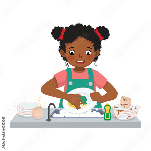 Cute little African girl washing dishes standing at sink in the kitchen doing housework chores at home