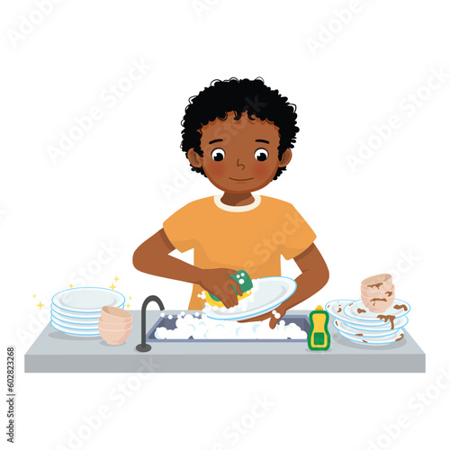 Cute little African boy washing dishes standing at sink in the kitchen doing housework chores at home