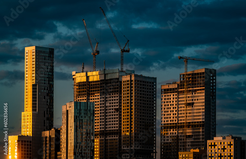 High-rise residential buildings under construction along with construction cranes illuminated by the setting sun against a dark blue sky
