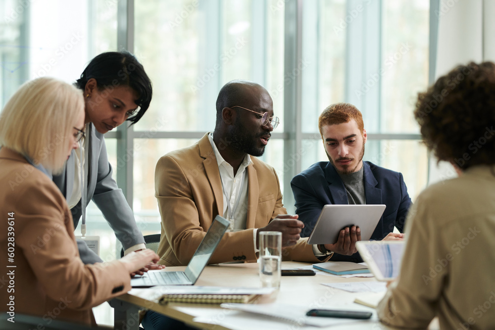 Confident analyst with tablet explaining online data to colleague during discussion of financial graphs and charts among coworkers in office Confident analyst with tablet explaining online data to colleague during discussion of financial graphs and charts among coworkers in office