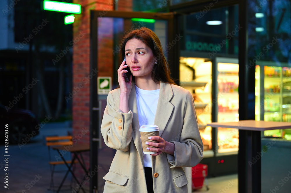 A beautiful woman dressed in casual attire is enjoying a cup of coffee in an urban setting. She appears to be deep in conversation as she speaks on her phone, with an expression