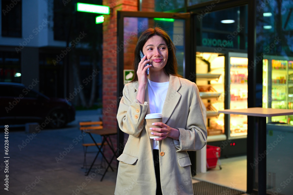 Fototapeta premium A beautiful woman dressed in casual attire is enjoying a cup of coffee in an urban setting. She appears to be deep in conversation as she speaks on her phone, with an expression