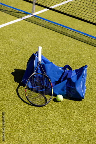 Tennis racket, ball and sports bag on the ground by net on sunny outdoor grass court, copy space