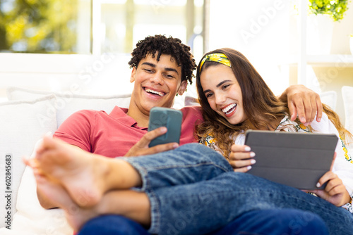 Happy diverse couple using smartphone and tablet relaxing on sofa in living room and laughing