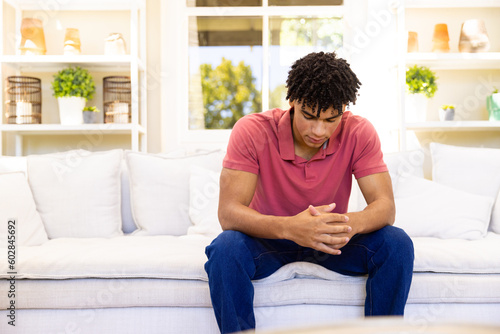 Worried, sad biracial man sitting on sofa in living room thinking, copy space