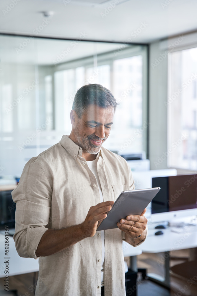 Vertical portrait Latin Hispanic mature adult professional business man, smiling Indian senior businessman CEO holding digital tablet using online finance application standing inside company office.