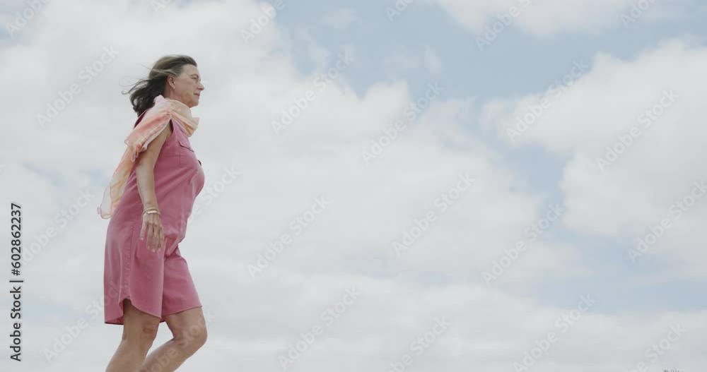 Happy senior caucasian woman walking against cloudy sky on beach and raising arms, in slow motion Happy senior caucasian woman walking against cloudy sky on beach and raising arms, in slow motion