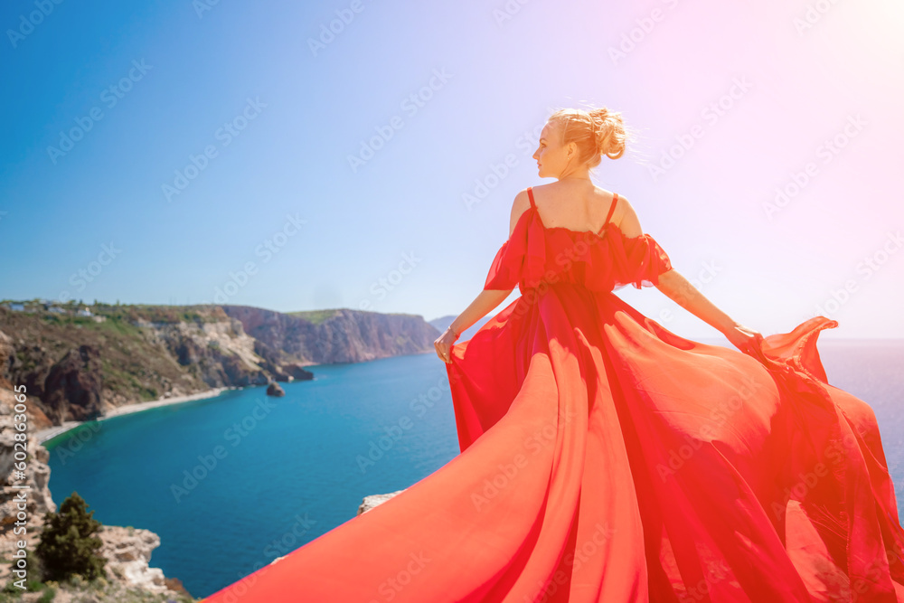 woman sea red dress. Blonde with long hair on a sunny seashore in a red flowing dress, back view, silk fabric waving in the wind. Against the backdrop of the blue sky and mountains on the seashore. woman sea red dress. Blonde with long hair on a sunny seashore in a red flowing dress, back view, silk fabric waving in the wind. Against the backdrop of the blue sky and mountains on the seashore.