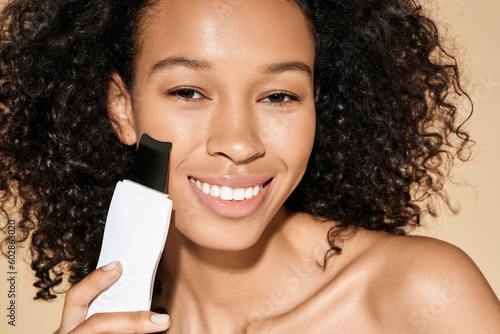 Fotografie African American woman with ultrasound scrubber treating her skin for remove blackheads and cleansing facial skin, on beige background