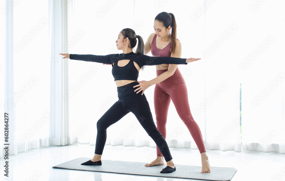 Naklejka premium Asian professional fitness trainer training teaching pretty healthy fit slim sexy female sporty athlete model in sports bra and leggings standing stretching legs balancing do warrior pose on yoga mat