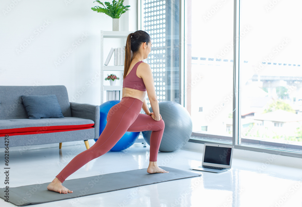 Asian beautiful healthy fit slim sexy female sporty athlete model in sports bra and leggings standing stretching legs on yoga mat warm up ready to workout exercise lesson via laptop in living room Asian beautiful healthy fit slim sexy female sporty athlete model in sports bra and leggings standing stretching legs on yoga mat warm up ready to workout exercise lesson via laptop in living room