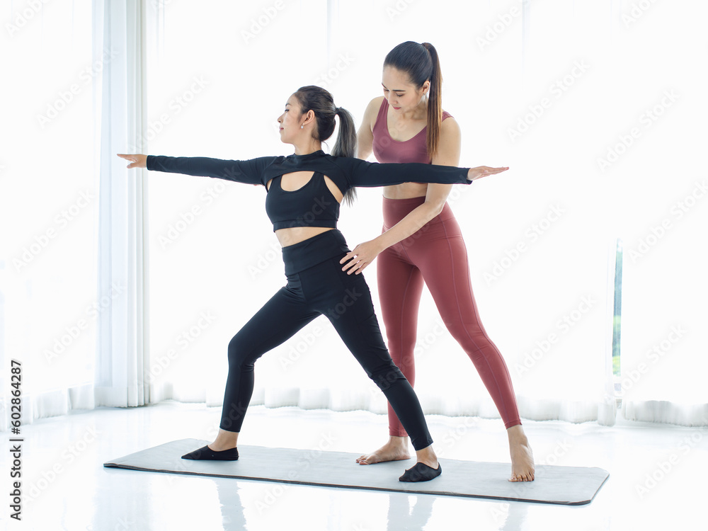 Asian professional fitness trainer training teaching pretty healthy fit slim sexy female sporty athlete model in sports bra and leggings standing stretching legs balancing do warrior pose on yoga mat