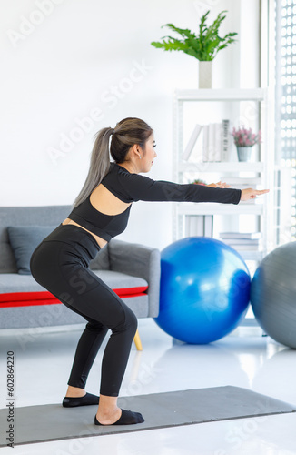 Asian beautiful healthy fit slim female sporty athlete model in black sports bra and leggings standing balancing squating on yoga mat ready to workout exercise in living room at home with rubber ball