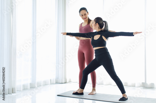 Asian professional fitness trainer training teaching pretty healthy fit slim sexy female sporty athlete model in sports bra and leggings standing stretching legs balancing do warrior pose on yoga mat