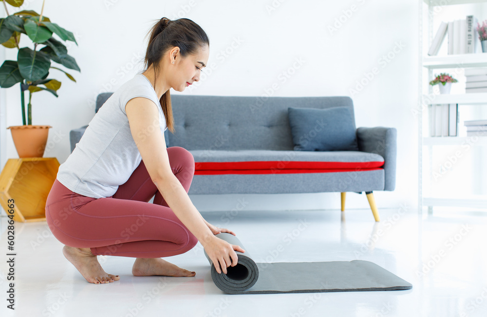 Fototapeta premium Asian beautiful healthy fit slim sexy female sporty athlete model in sportswear and leggings sitting bending head down rolling yoga mat out ready for workout exercising training in living room
