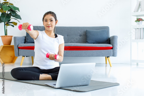 Portrait shot Asian beautiful healthy fit slim sexy female sporty athlete model in sportswear and leggings sitting smiling look at camera lifting up dumbbells workout exercising via laptop at home